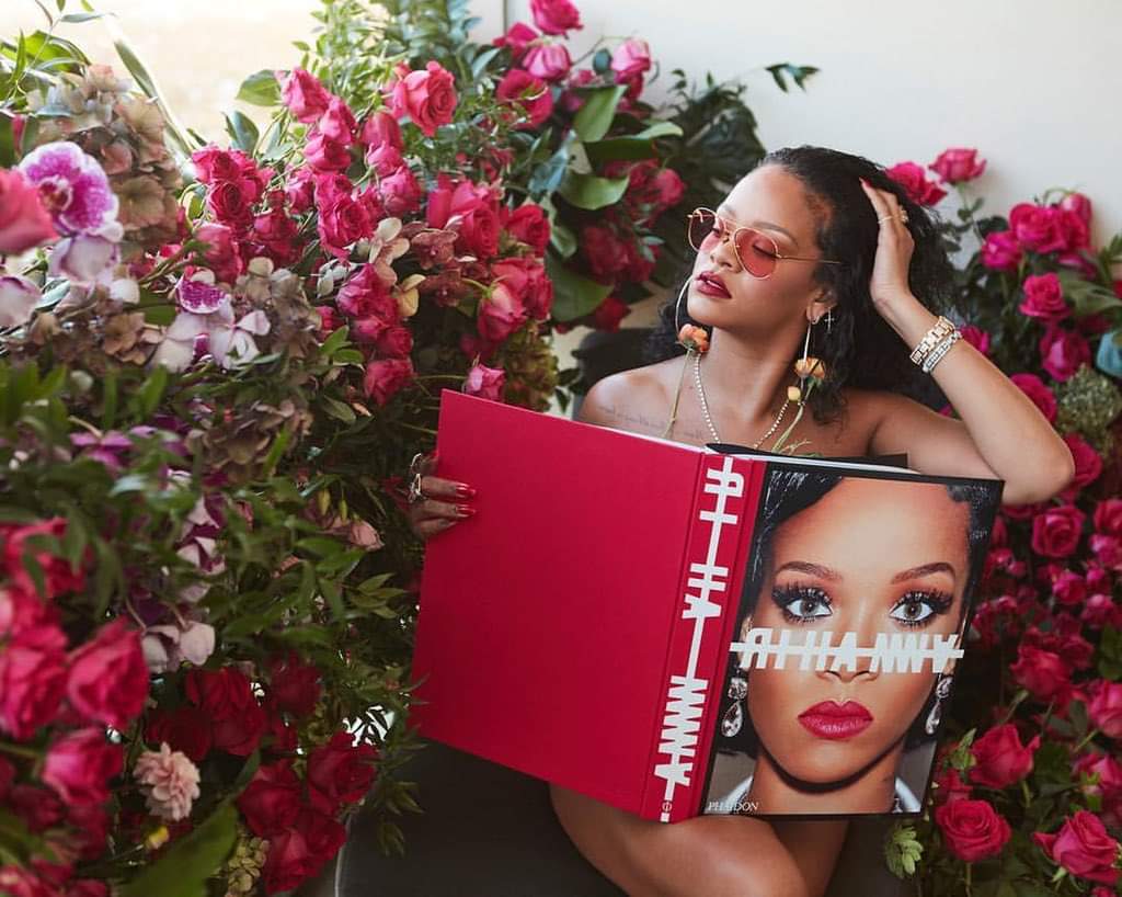 Cantora Rhianna posa sentada com um livro vermelho sob seu colo ao lado de muitas rosas