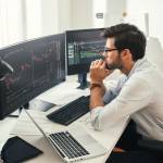 Successful traders. Back view of bearded stock market broker in eyeglasses analyzing data and graphs on multiple computer screens while sitting in modern office. Master Trader