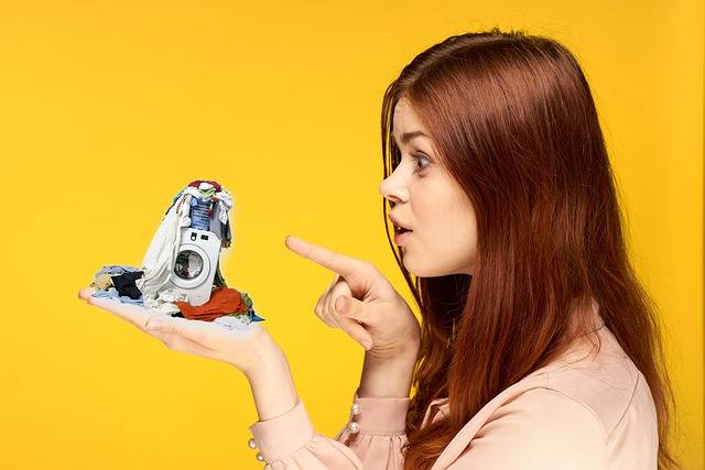Montagem de uma moça ruiva em um fundo amarelo segurando uma mini máquina de lavar roupas na mão esquerda e apontando para ela com a direita