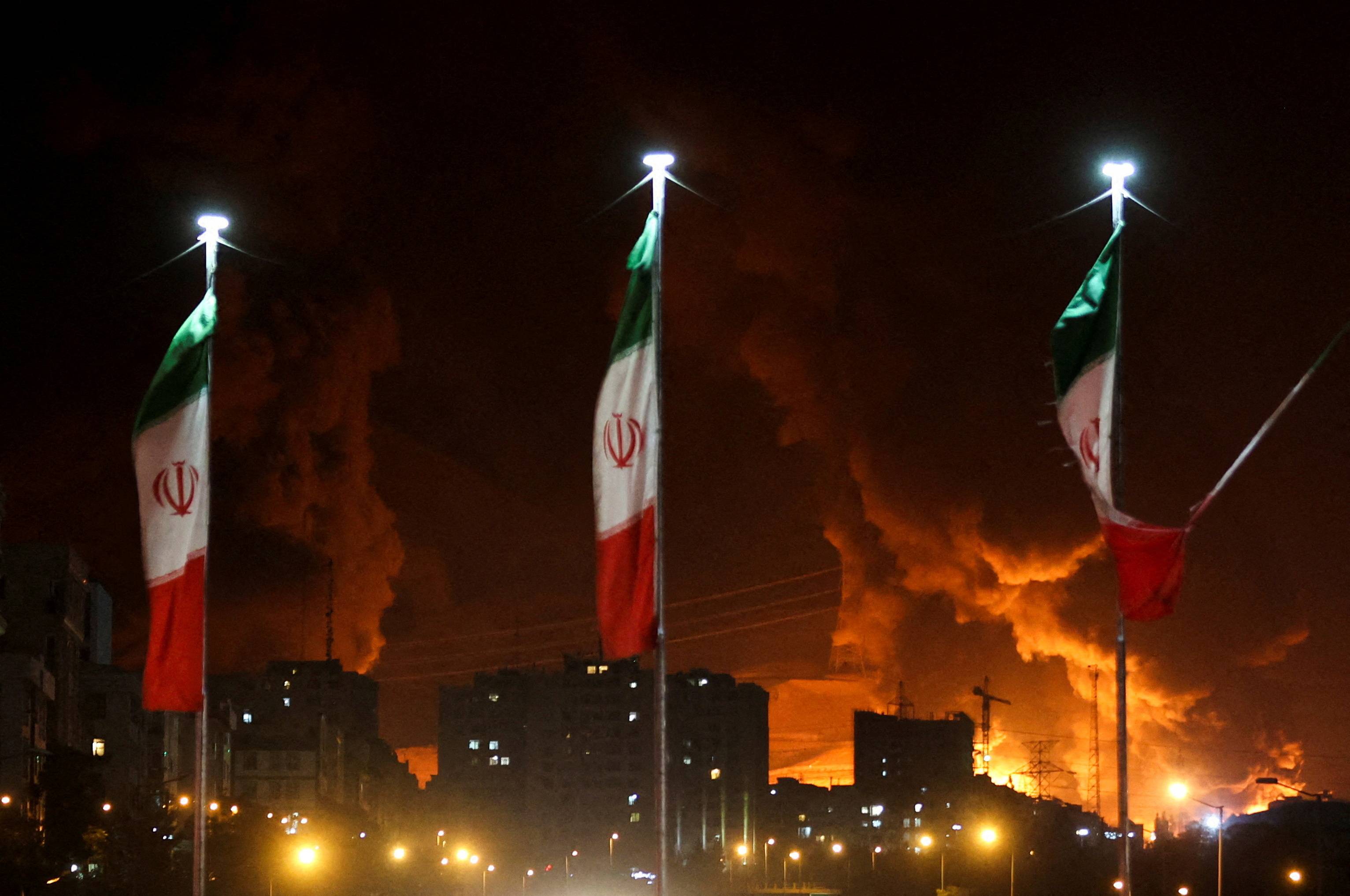 Bandeiras iranianas tremulam enquanto fogo e fumaça de um ataque israelense ao depósito de petróleo de Sharan aumentam, após ataques israelenses ao Irã, em Teerã, Irã, 15 de junho de 2025. Majid Asgaripour/WANA (West Asia News Agency) via REUTERS