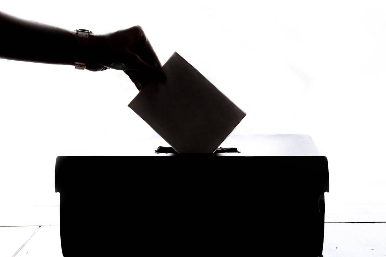 A imagem mostra uma silhueta de mão colocando um voto na urna; crédito: imagem de banco livre.